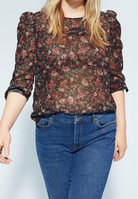 Durchsichtige schwarze Bluse mit einem floralen Muster in Rot und Grün. Sie hat Puffärmel und einen lockeren Schnitt und wird mit blauen Jeans kombiniert.