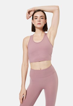 Młoda kobieta w wrzosowym krótkim topie i legginsach rozciąga się z uniesioną jedną ręką na jednolitym tle.