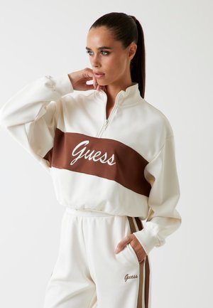 Felpa zip-up crema con striscia orizzontale marrone e logo ricamato "Guess", abbinata a pantaloni jogger crema con accenti laterali marroni.