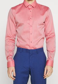 Uomo che indossa una camicia button-up a maniche lunghe corallo, aderente, infilata in pantaloni blu su misura.
