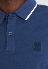 Mørkeblå polo-shirt med struktureret stof, der har en hvid krave-stribe og et præget firkantet mærke med teksten "BOSS." Tre knaplukninger.