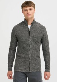 Cárdigan gris de cremallera con cuello alto, que presenta un patrón de punto texturizado y mangas largas, llevado sobre una camiseta blanca.