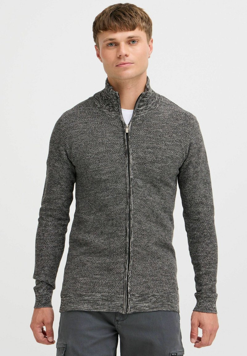 Cárdigan gris de cremallera con cuello alto, que presenta un patrón de punto texturizado y mangas largas, llevado sobre una camiseta blanca.