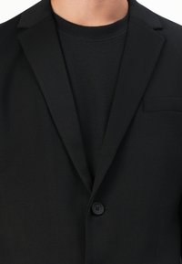 Sort blazer med skrå krave, enkelt knaplukning og åben brystlomme, lagret over en struktureret sort skjorte. Glat stoffinish.