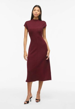 Naine kannab keha järgivat, varrukateta burgundia midi-kleiti kõrge kaelusega ja musti avatud varbakärgiga kontsakingi, seisab valge tausta ees.