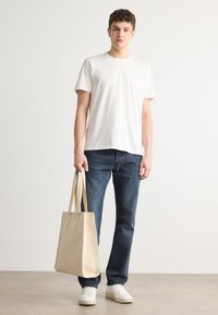 White cotton t-shirt, blue denim jeans, and beige canvas tote bag. Model wears white sneakers. Smooth texture, simple design.