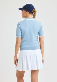 Jasnoniebieska, krótko rękawowa koszulka polo z białym kołnierzykiem i mankietami, w połączeniu z białą plisowaną spódnicą. Materiał wydaje się dzianinowy i gładki. Nosi granatową czapkę.