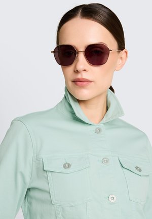 Femme aux cheveux foncés portant des lunettes de soleil angulaires foncées et une veste vert menthe clair à col relevé sur un fond pâle.