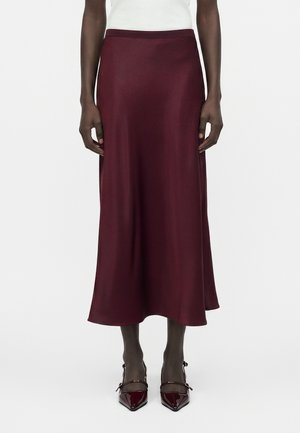 Žena nosiaca bordovú midi sukňu spojenú s rovnako sfarbenými lodičkami a bielym topom, stojaca pred jednofarebným pozadím.