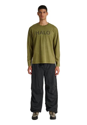 Junger Mann trägt ein olivgrünes Langarmshirt mit dem Aufdruck "HALO", schwarze weite Hose und Wanderschuhe, steht vor einem weißen Hintergrund.