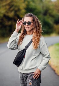 Světle šedá oversized mikina, černá ledvinka, zubožené šedé džíny, kulaté sluneční brýle, vlnité vlasy po ramena, venku na cestě.