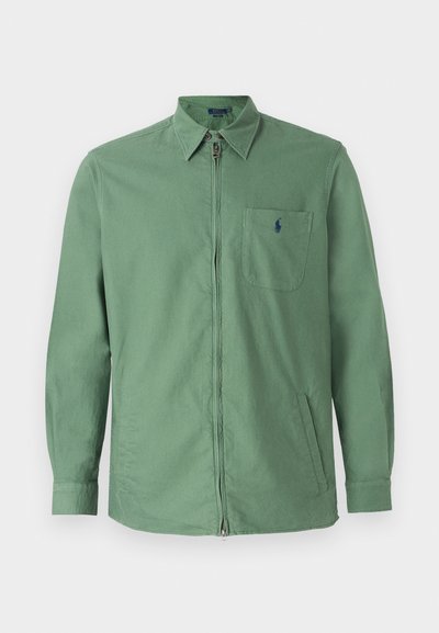 Chaqueta de algodón verde con cierre frontal, cuello abotonado y un bolsillo en el pecho con un logo. Corte recto y mangas largas.