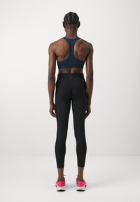 Sötétkék sportmelltartó, amely versenyhátas dizájnnal rendelkezik. Feszes fekete leggings sima textúrával, rózsaszín sportcipőkkel párosítva.