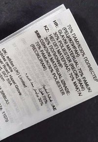 Etichetta con più lingue; elenca i materiali: 70% cotone, 30% poliestere. Sfondo testurizzato argentato con dettagli stampati neri.