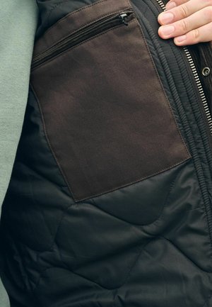 Zwarte gewatteerde jas interieur met een bruine stoffen zak en ritssluiting. Textuur met zichtbare stiksels en ritsdetails.