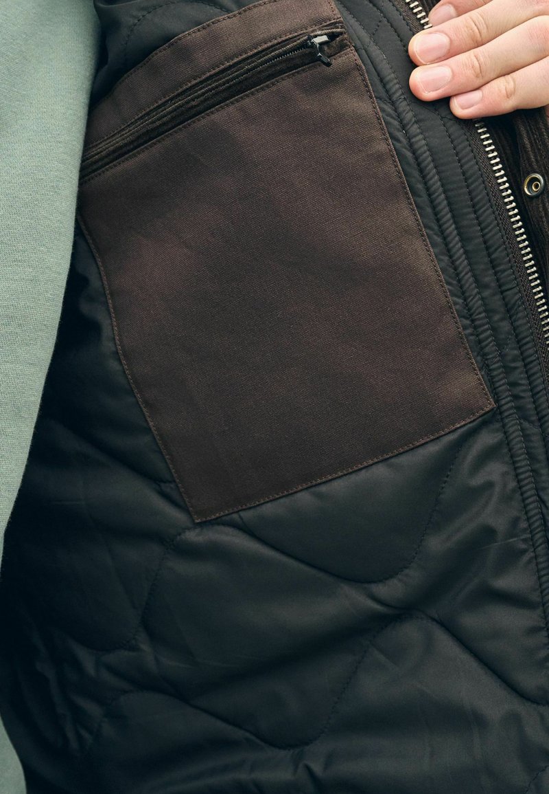 Interior de chaqueta negra acolchada con un bolsillo de tela marrón y cierre de cremallera. Superficie texturizada con costuras visibles y detalle de cremallera.