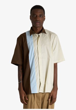 Giovane uomo in piedi, che indossa una camicia a maniche corte con pannelli verticali marroni, beige e azzurri, e pantaloni beige.