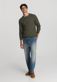 Maglione a coste verde oliva con scollatura a girocollo, abbinato a jeans azzurri a gamba affusolata e scarpe marroni con lacci.