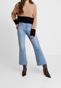 Femme portant un pull en maille beige et noir, un jean flare bleu clair raccourci, et des bottes pointues en imprimé serpent marron foncé, debout avec la main sur la hanche.