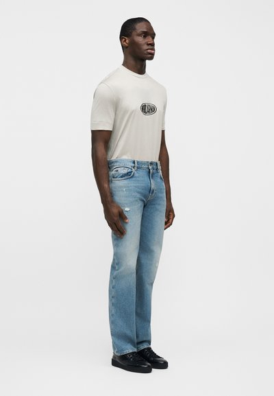 Μπεζ κοντομάνικη μπλούζα με μαύρο λογότυπο, συνδυασμένη με ανοιχτό μπλε τζιν παντελόνι με ίσια γραμμή. Το μοντέλο φοράει μαύρα αθλητικά παπούτσια. Μινιμαλιστικός σχεδιασμός, εφαρμοστή γραμμή.