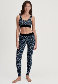 Vrouw die blootsvoets staat in een bijpassende zwarte sportbeha en legging met blauw-witte bloemenprint tegen een lichte achtergrond.