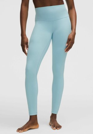 Persoon die lichtblauwe leggings met hoge taille draagt, staat blootsvoets op een effen lichte achtergrond.