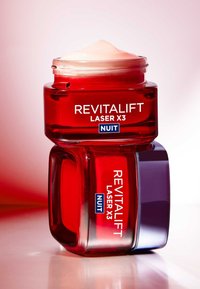 Bocal en verre rouge avec un couvercle en crème rose, étiqueté "REVITALIFT LASER X3" et "NUIT". Le bocal a une forme carrée et une texture lisse.