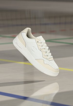 Hvit og beige lavsko med lisser, svevende over et gymsalgulv merket med fargede linjer.