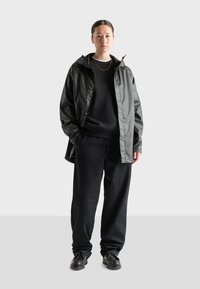 Chaqueta negra impermeable con capucha sobre un suéter negro, combinada con pantalones negros holgados y zapatos negros. Diseño simple y funcional.