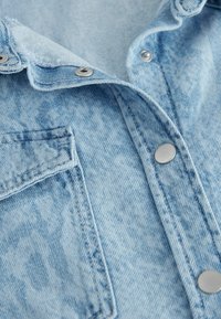 Camicia di jeans azzurro chiaro con colletto, bottoni a pressione argentati e taschino sul petto. Il tessuto ha una finitura testurizzata con leggere variazioni.