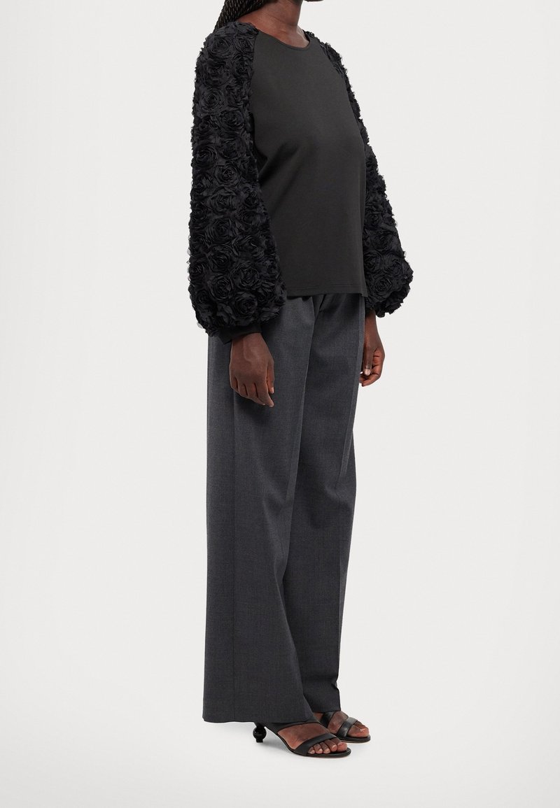 Haut noir avec des manches bouffantes ornées de fleurs ; associé à un pantalon taille haute gris et des sandales noires à bout ouvert.