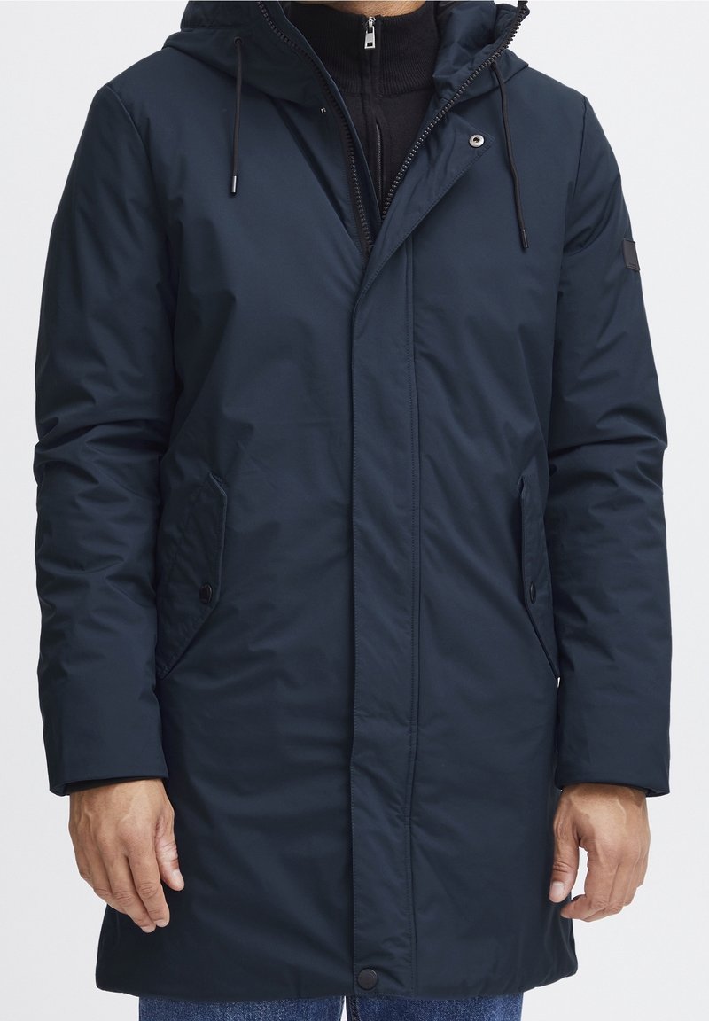 Parka bleu marine avec fermeture à glissière et pressions, capuche ajustable, poches latérales et texture lisse résistante à l'eau. Présente un logo sur la manche.