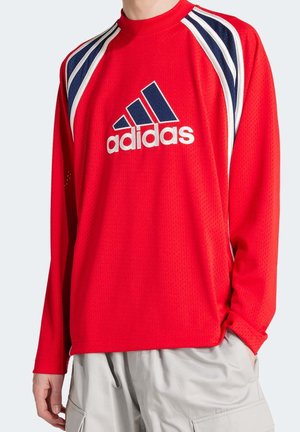 Personne portant un maillot de sport Adidas rouge à manches longues avec des bandes bleu marine et blanches sur les épaules et un pantalon cargo gris clair.