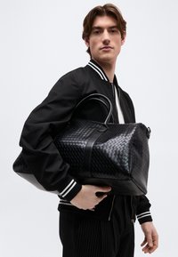 Jeune homme en veste bomber noire tenant un grand sac de voyage en cuir tressé noir sur fond blanc.