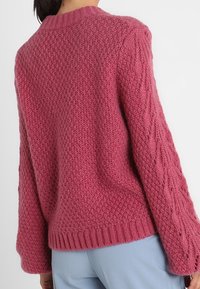 Pull en grosse maille rose avec un motif texturé, ourlet côtelé et détails en tricot torsadé sur les manches. Coupe confortable et ample.
