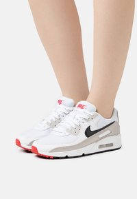 Vita nät-sneakers med grå och svarta accenter, featuring röda detaljer på sulan och tungan. Inkluderar traditionell snörning och synlig luftenhet.