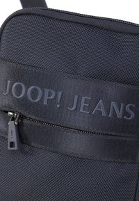 Mjärksblå tyg axelväska med texturerad yta, en dragkedjeficka och präglad "JOOP! JEANS" märkning på framsidan.