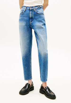 Osoba nosząca jasnoniebieskie dżinsy z wysokim stanem z wyblakłymi detalami i czarne skórzane mokasyny, stojąca na białym tle.