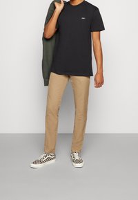 Svart t-shirt i bomull med "OBEY"-logotyp, beige chinos och svarta och vita rutiga sneakers. Modellen håller en grön jacka upphängd över ena axeln.