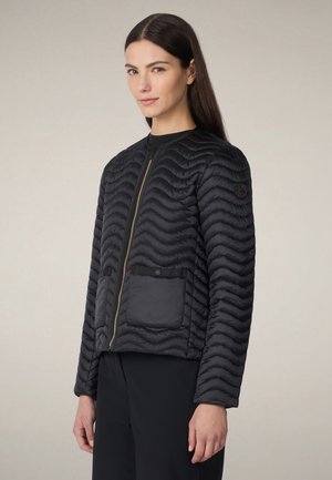 Frau trägt eine schwarze gesteppte Jacke mit Frontreißverschluss, einer Druckknopftasche und langen Ärmeln, kombiniert mit schwarzen Hosen.