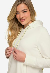 Felpa bianca con cappuccio, dotata di un cordino con dettagli verde chiaro, polsini a coste e un logo vicino alla spalla. Tessuto morbido.