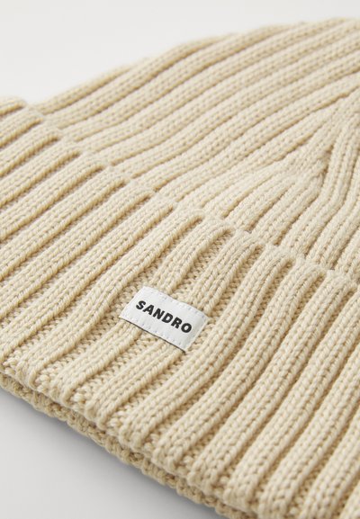 Šviesiai pilka megzta kepurė su vertikaliomis raukšlėmis, kurioje yra balta etiketė su "SANDRO" juoda spalva. Minkšta tekstūra ir tvirtas prigludimas.