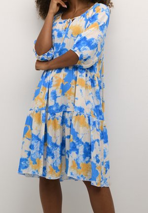 Femme portant une robe batik bleu, blanc et jaune jusqu'aux genoux, avec des manches jusqu'aux coudes, debout les bras croisés et la main sur le menton.