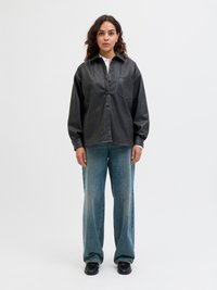 Black faux leather button-up shirt with a chest pocket, paired with wide denim pants and black shoes. Model stands against a white background.