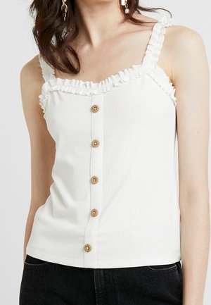 Kobieta ubrana w biały, bezrękawnik z prążkowanego materiału z falbanianymi ramiączkami i drewnianymi guzikami z przodu, w połączeniu z czarnymi spodniami.