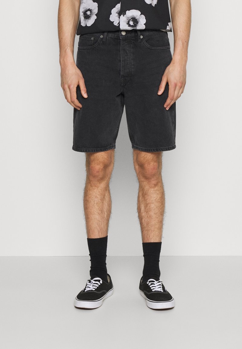Schwarze Jeansshorts mit geradem Schnitt, ausgestattet mit fünf Taschen und Gürtelschlaufen. Getragen mit schwarzen Socken und Leinenschuhen.