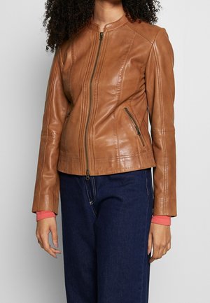 Femme portant une veste en cuir marron cintrée avec une fermeture éclair devant et des poches zippées, associée à un jean bleu foncé et un soupçon d'une manche de chemise rouge.
