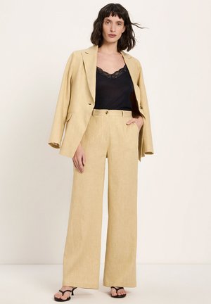 Woman wearing beige wide-leg suit with blazer draped over shoulders, black lace-trimmed camisole, and black heeled sandals standing against plain background.