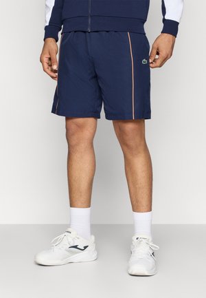 Un mannequin porte un short de sport bleu marine avec un passepoil orange et un petit logo crocodile, des chaussettes blanches et des baskets blanches.