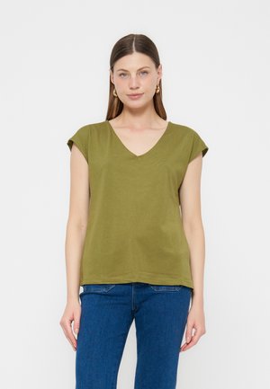 Woman with straight brown hair wears green short-sleeve V-neck top and blue jeans, standing against plain white background.